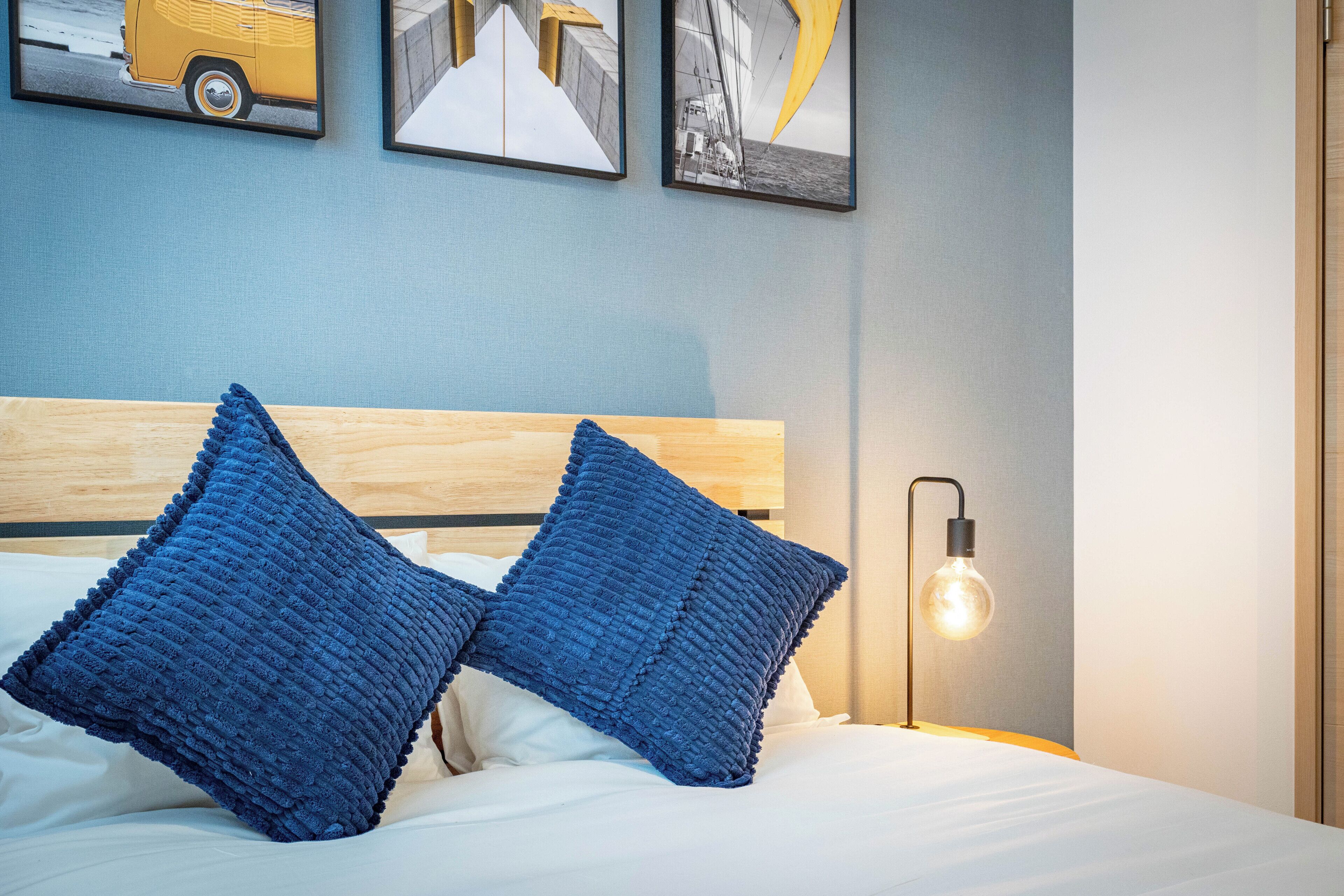 Cama bem arrumada com duas almofadas azuis e cabeceira de madeira. Há um candeeiro fixado na parede e três quadros emoldurados sobre a cama.