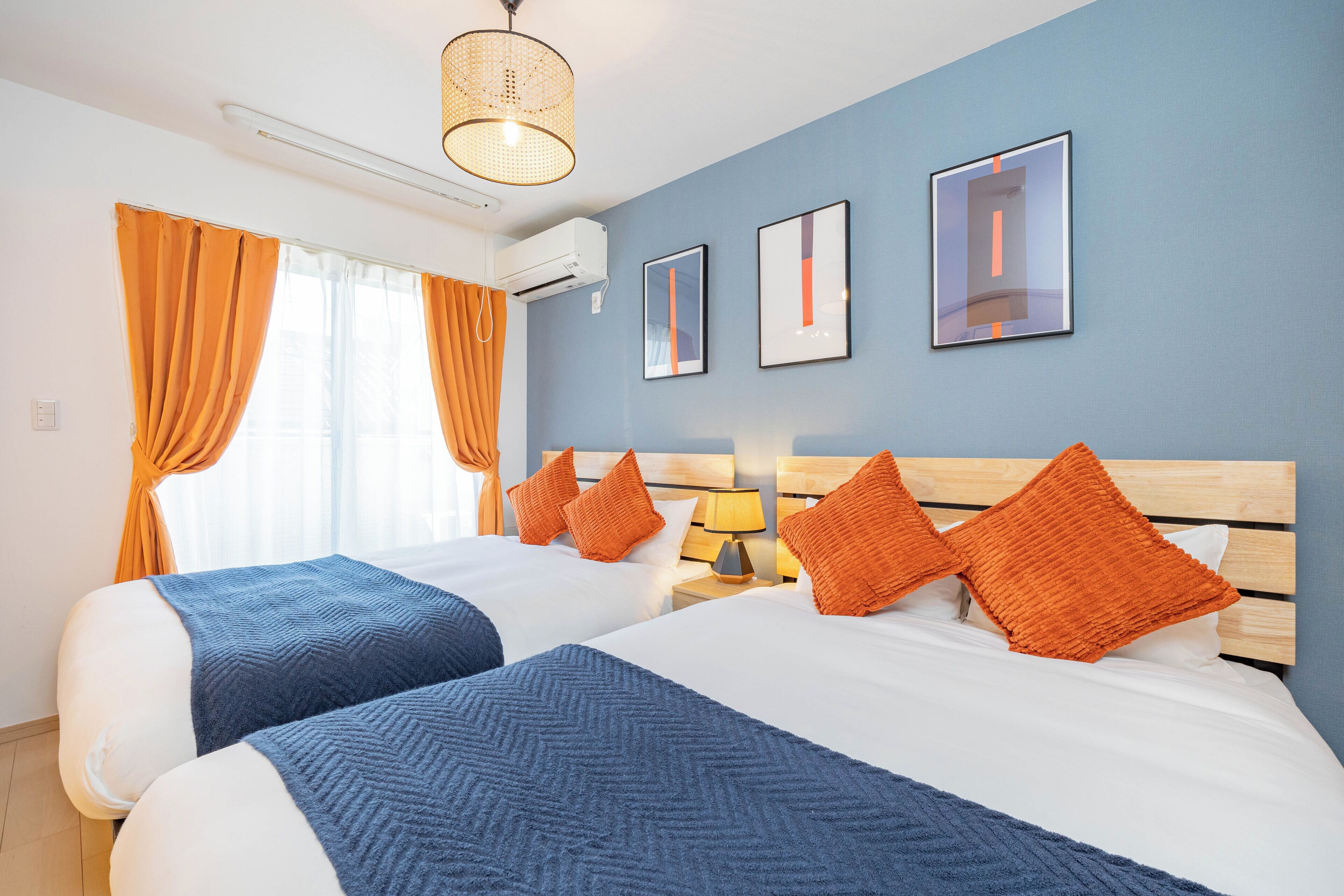 A hotel room with two beds, orange curtains, a wooden headboard, a wall-mounted air conditioner, and framed pictures.