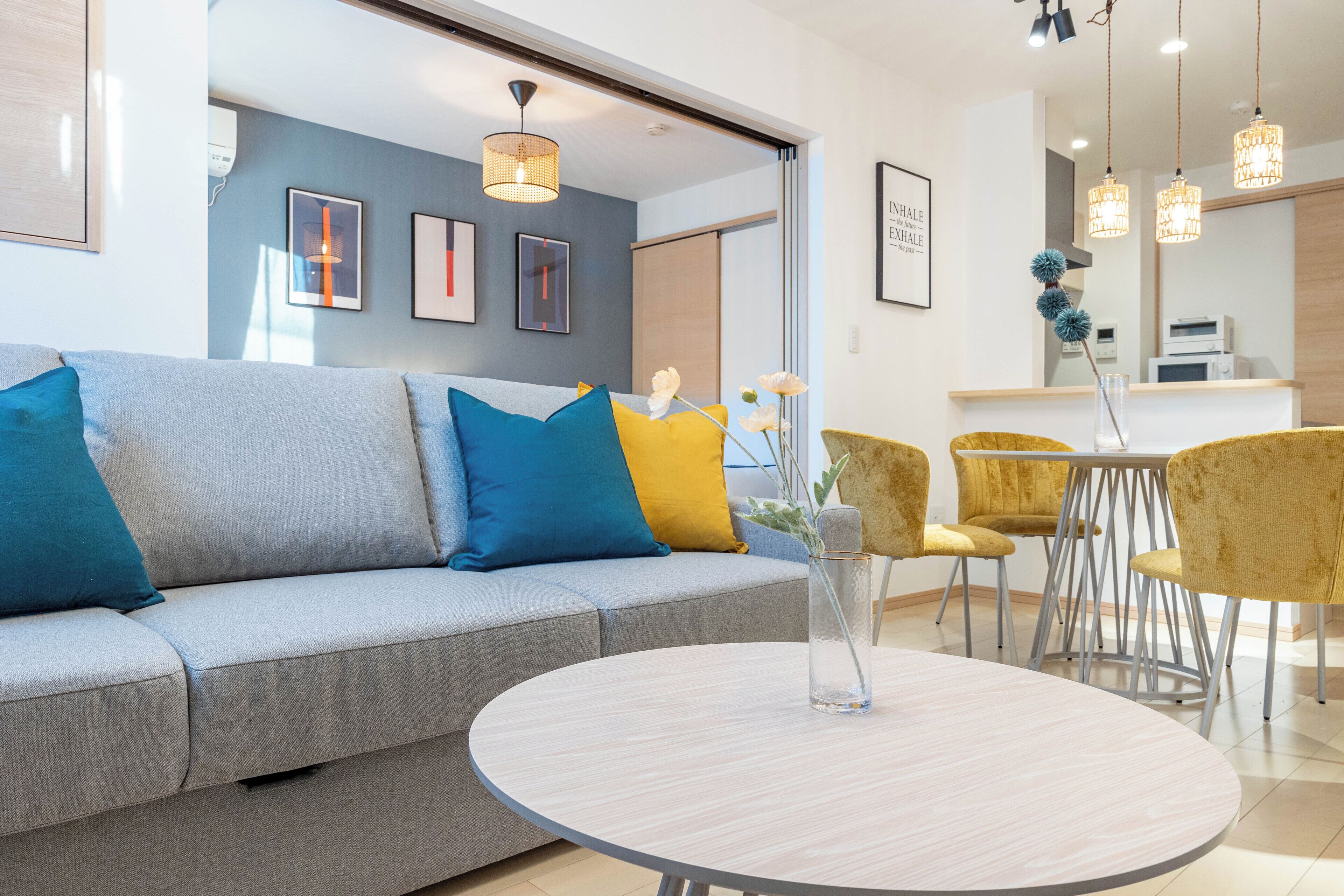 Una sala moderna con un sofá gris, cojines amarillos y azules, una mesa de centro redonda y un comedor con sillas amarillas.