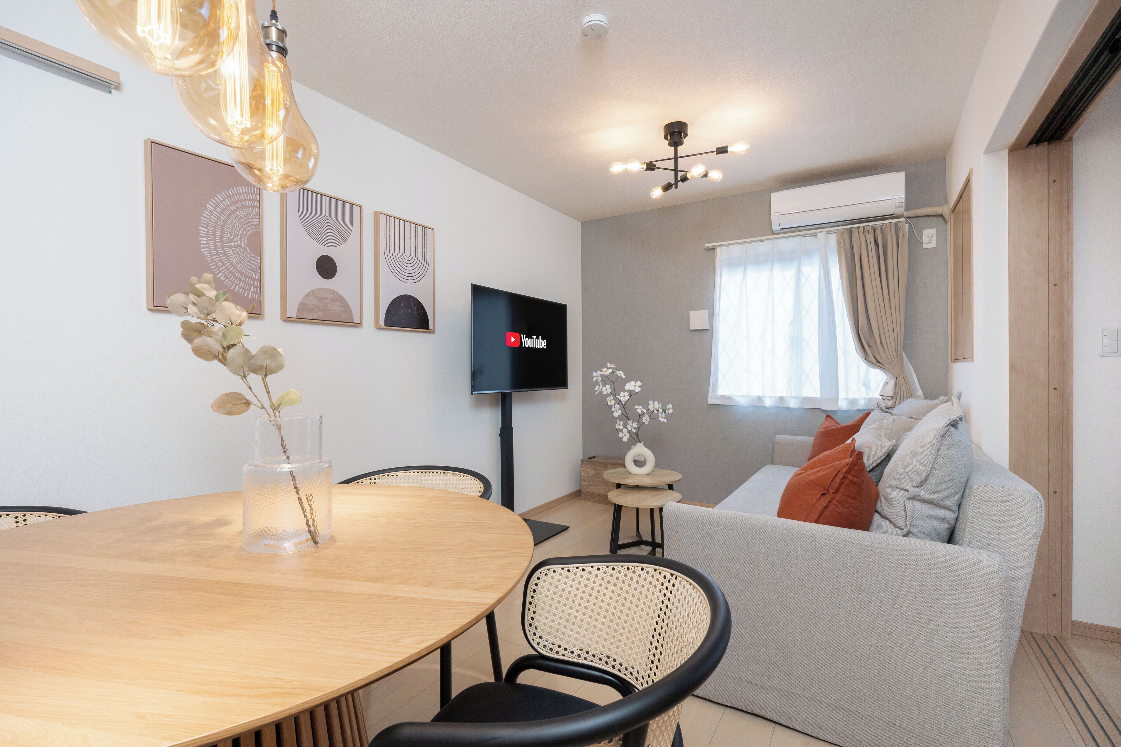 A modern living room with a round wooden dining table, a grey sofa with orange cushions, a flat-screen TV, and a vase with flowers.