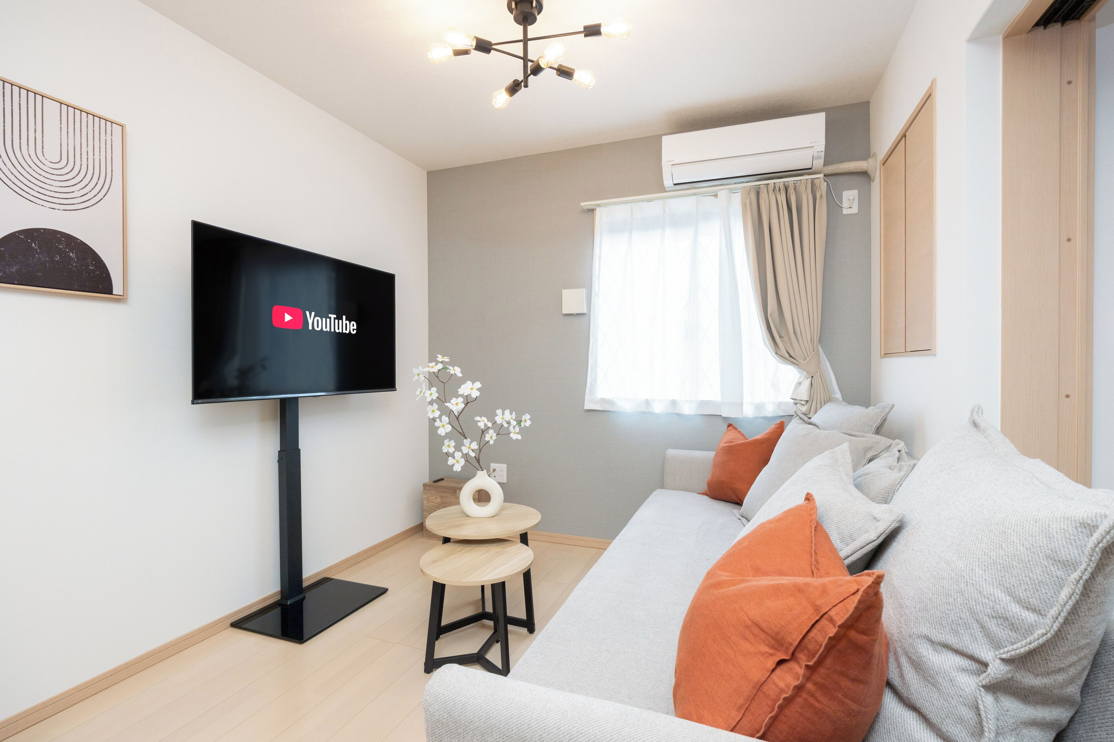 A modern living room with a flat-screen TV, a small round table, a sofa with orange cushions, a decorative vase, and a window with curtains.