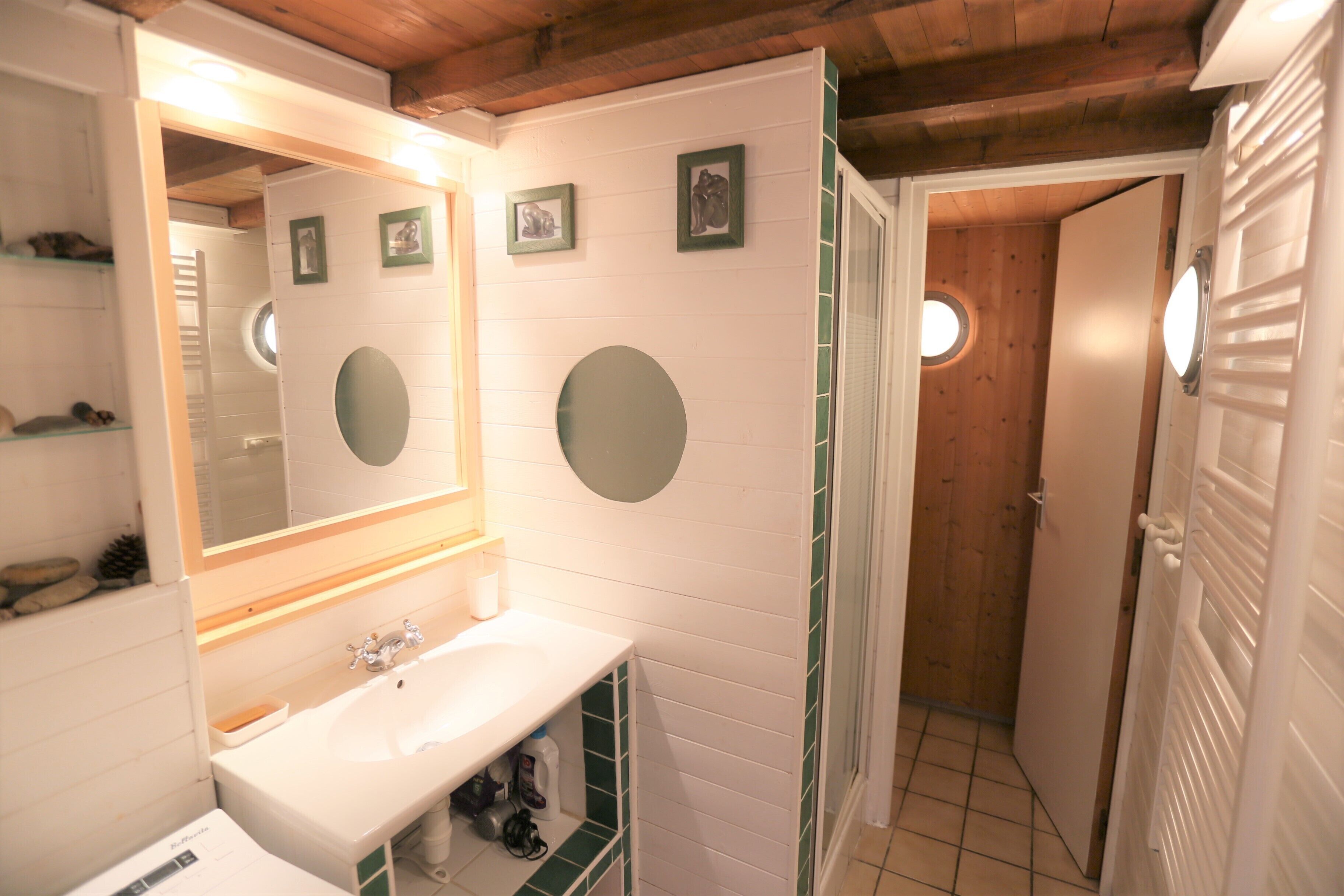 A bathroom with a white sink, mirror, and green-tiled walls. There are three framed pictures on the wall and a wooden door leading to another room.