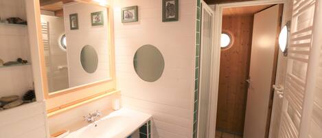 A bathroom with a white sink, mirror, and green-tiled walls. There are three framed pictures on the wall and a wooden door leading to another room.