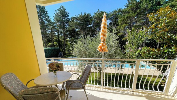 Een balkon met uitzicht op een zwembad, twee rieten stoelen, een kleine tafel met een vaas en een gestreepte parasol.