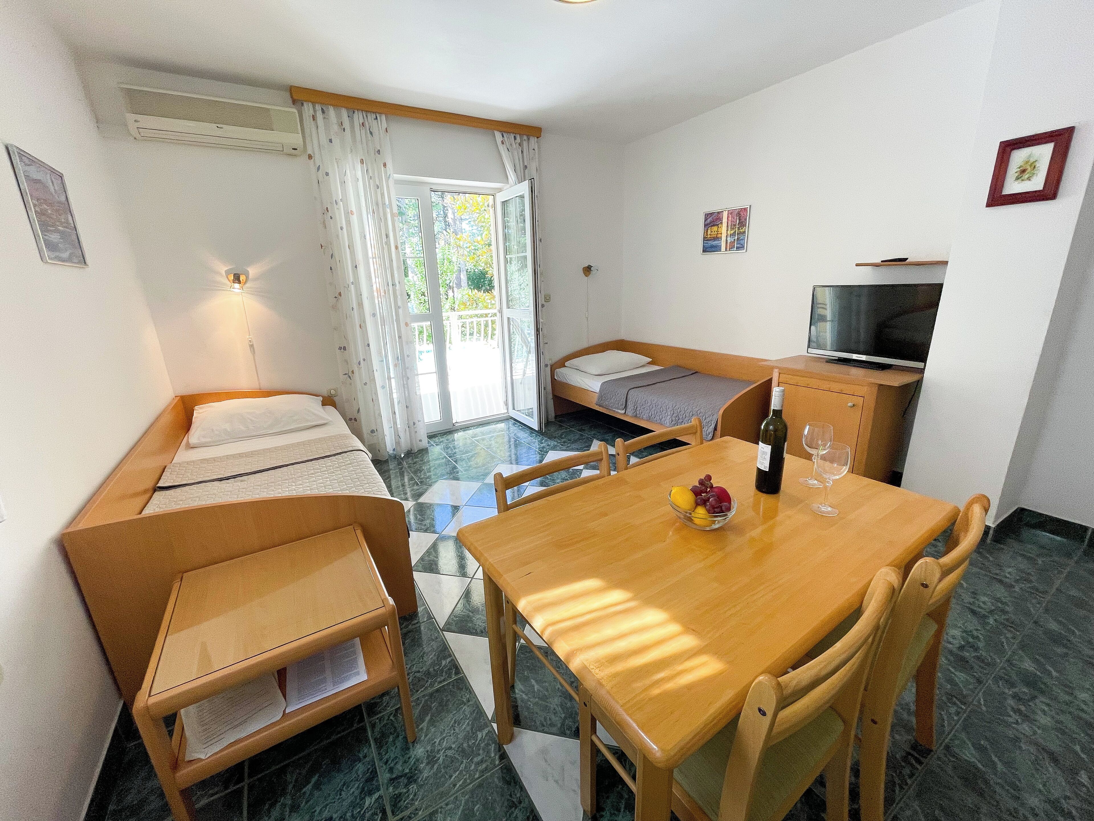 A compact room with a bed, a small sofa, a dining table with chairs, a television, and a balcony with curtains.