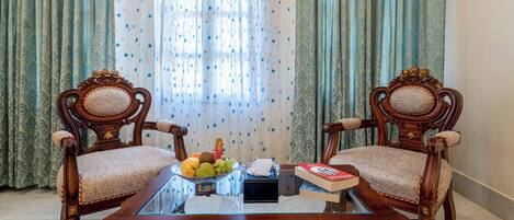 Una habitación con dos sillas de madera decoradas, una mesa de centro de vidrio con un frutero, un libro y una caja de pañuelos.