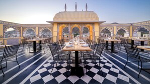 A rooftop dining area with checkered flooring, tables set for dining, and a view of a traditional building with arches. - BRIJ CASTLE (Jaipur)