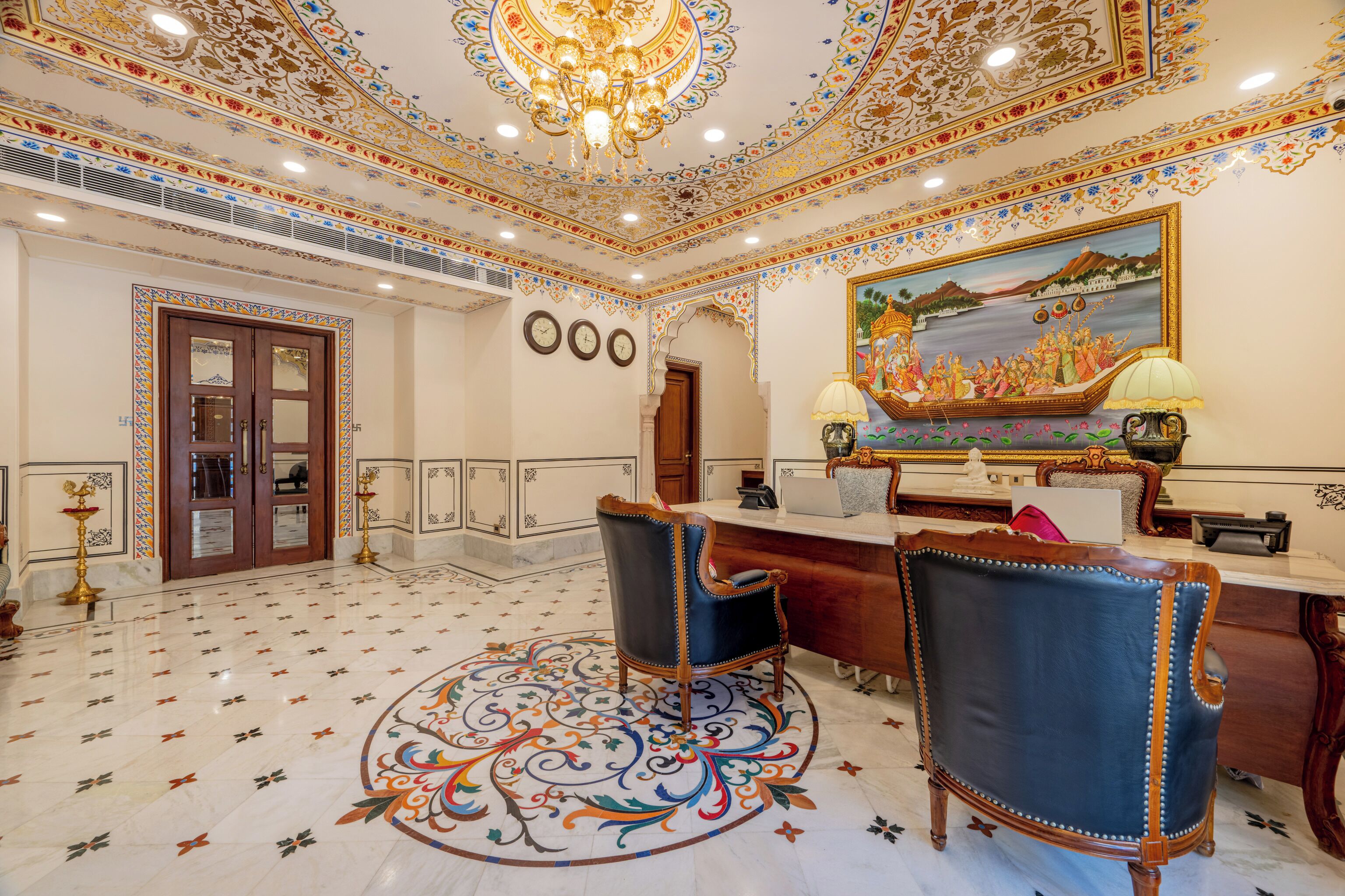 A luxurious lobby with ornate ceiling designs, a reception desk, two leather chairs, and a large decorative painting.