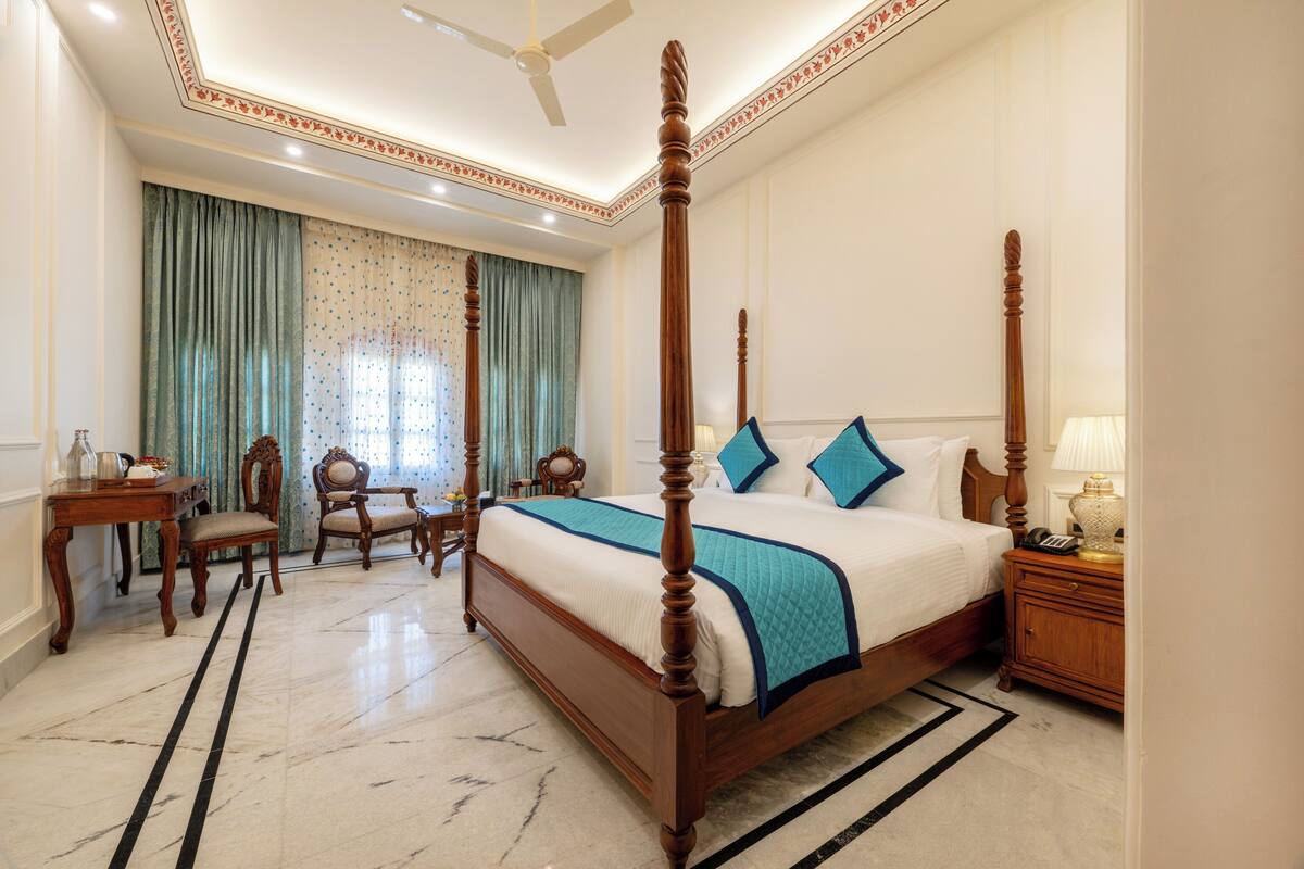 A spacious bedroom with a four-poster bed, a sitting area with a chair and table, and a large window with curtains.