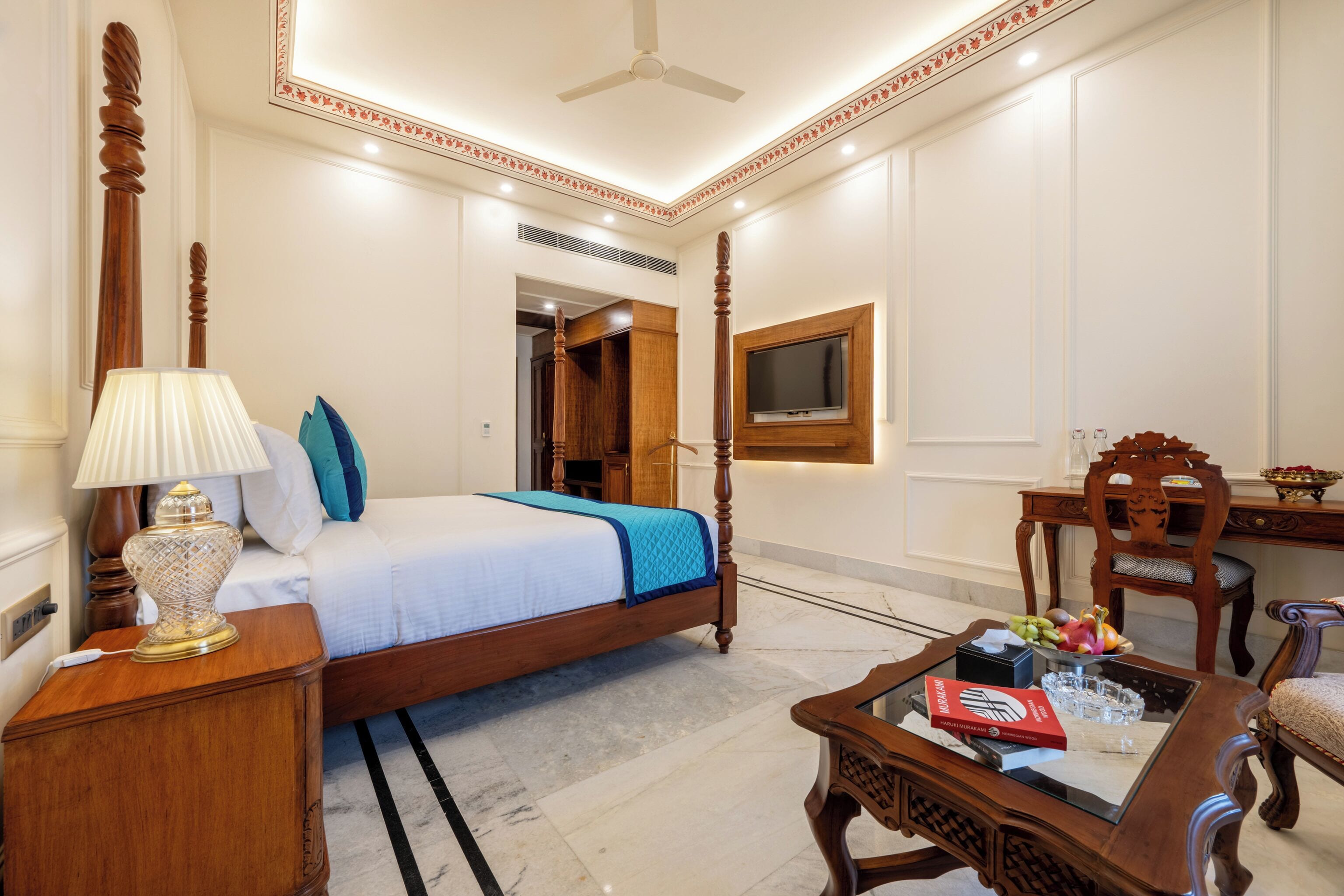 Habitación de hotel con cama con dosel, mesita de noche con lámpara, una silla de madera, escritorio con televisor y mesa de comedor con fruta y una caja roja.