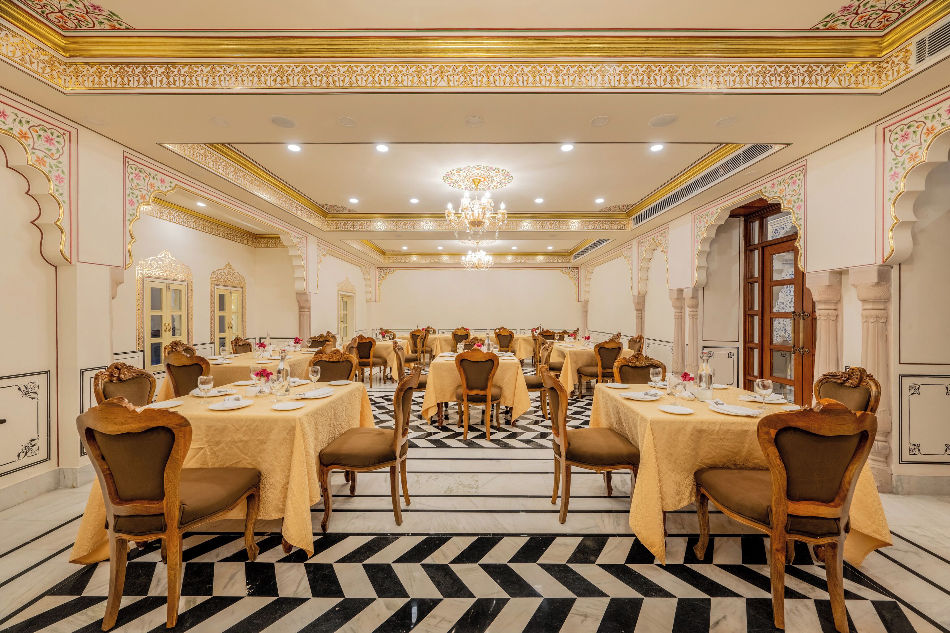 A formal dining room with multiple tables and chairs, ornate ceiling decorations, and a chandelier.
