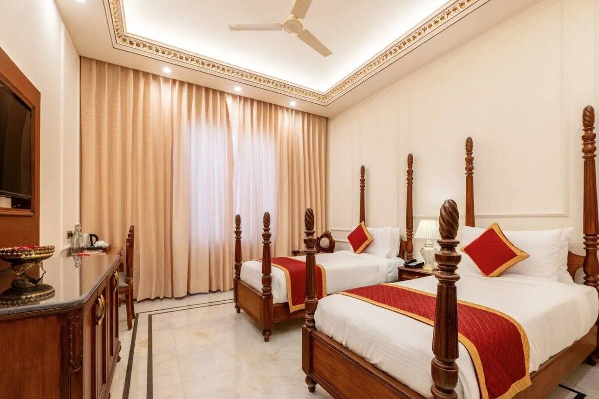 A hotel room with two wooden four-poster beds, a flat-screen TV, a desk with a lamp, and a large window with curtains.