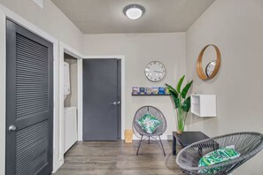 Un couloir avec une porte fermée, une horloge murale, une étagère remplie de livres, une plante en pot et un fauteuil en osier.