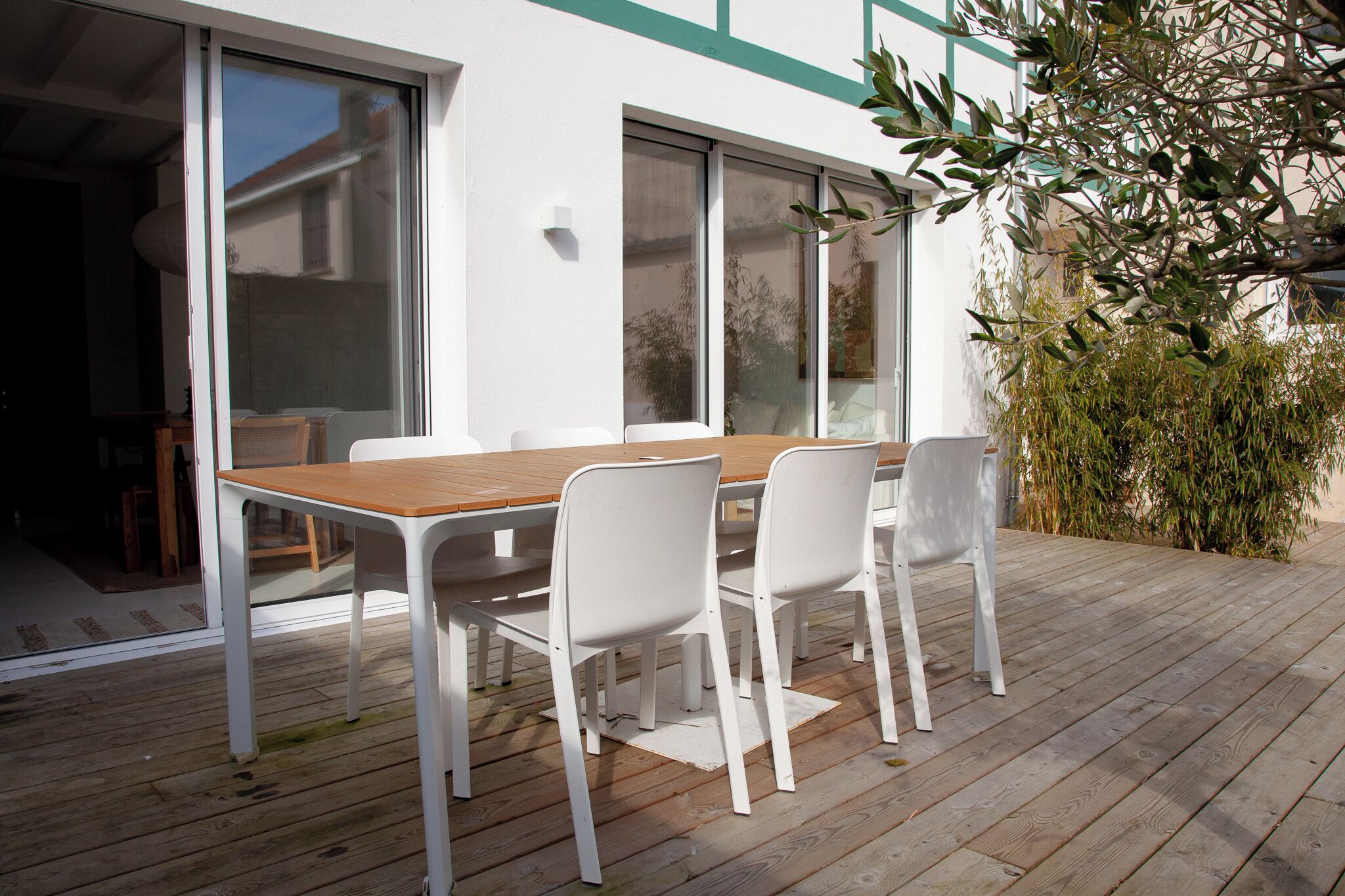 Une terrasse en bois avec une table et des chaises, de grandes portes-fenêtres coulissantes et une vue sur une maison au loin.