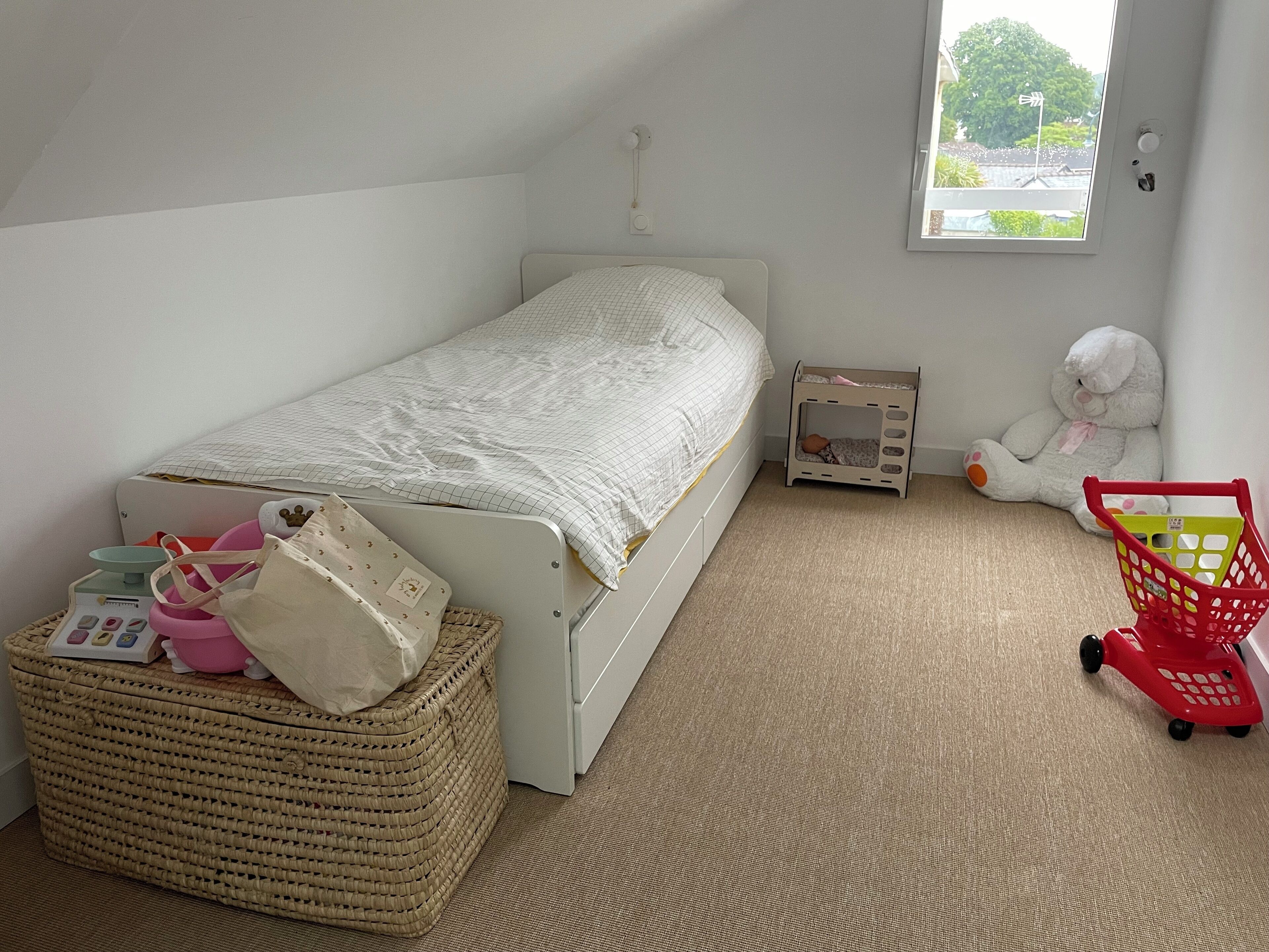 Un dormitorio pequeño y ordenado con una cama individual, una ventana, un carrito de juguete y un peluche.