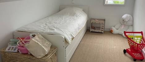 Un dormitorio pequeño y ordenado con una cama individual, una ventana, un carrito de juguete y un peluche.