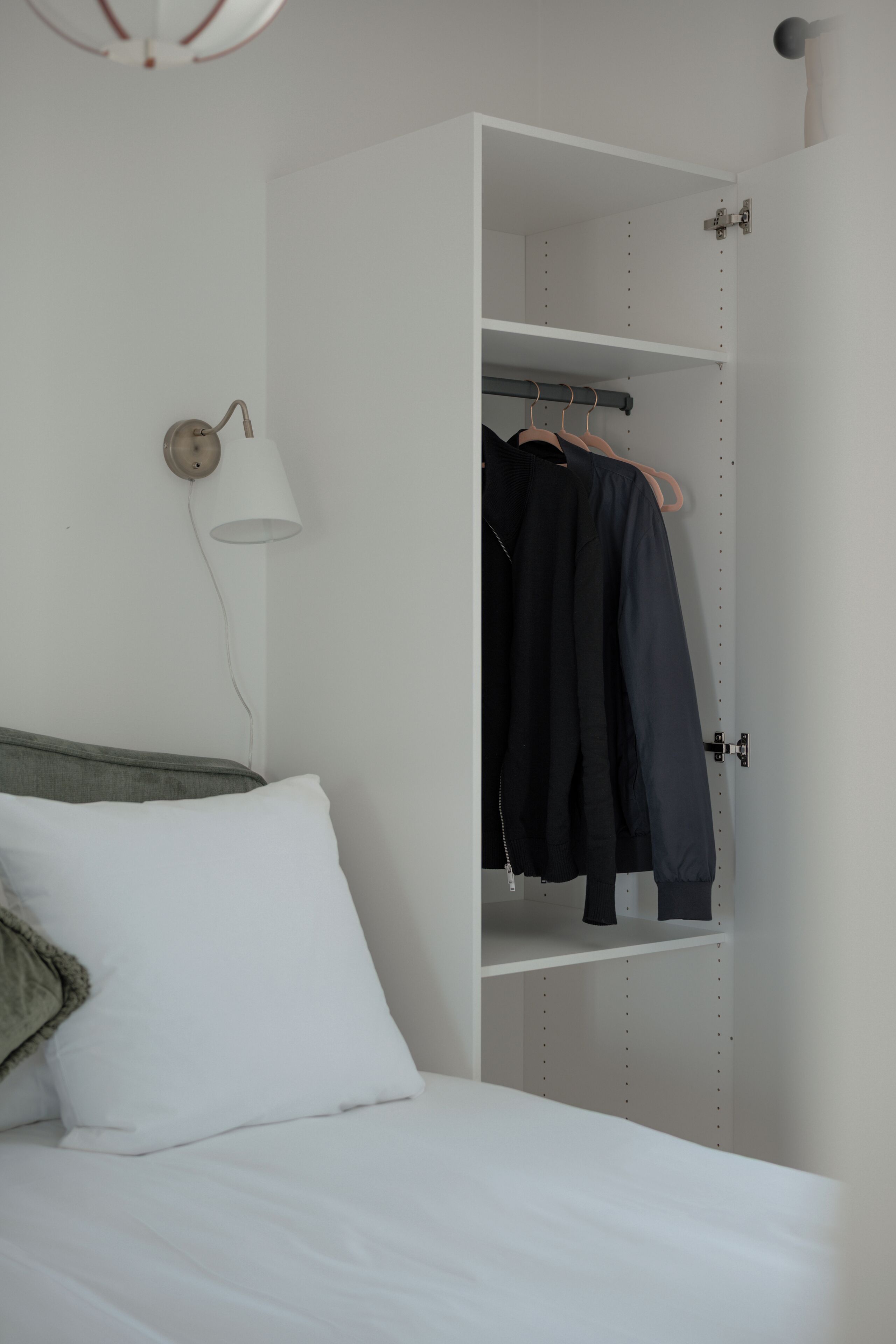 Une chambre à coucher avec un lit, un mur blanc, une lampe suspendue et une armoire ouverte remplie de vêtements.