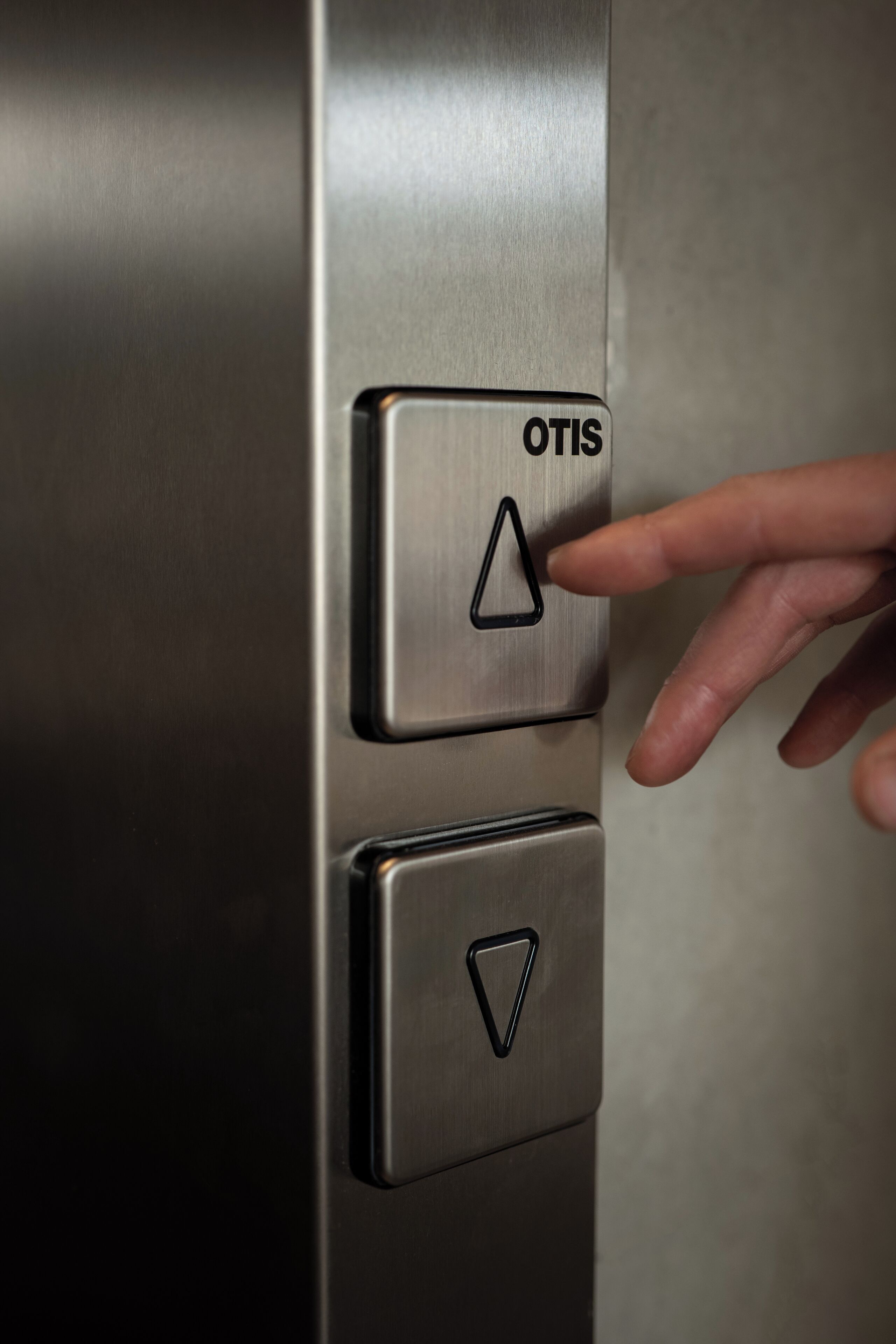 A metallic elevator button with the label "OTIS" and a hand pressing it.