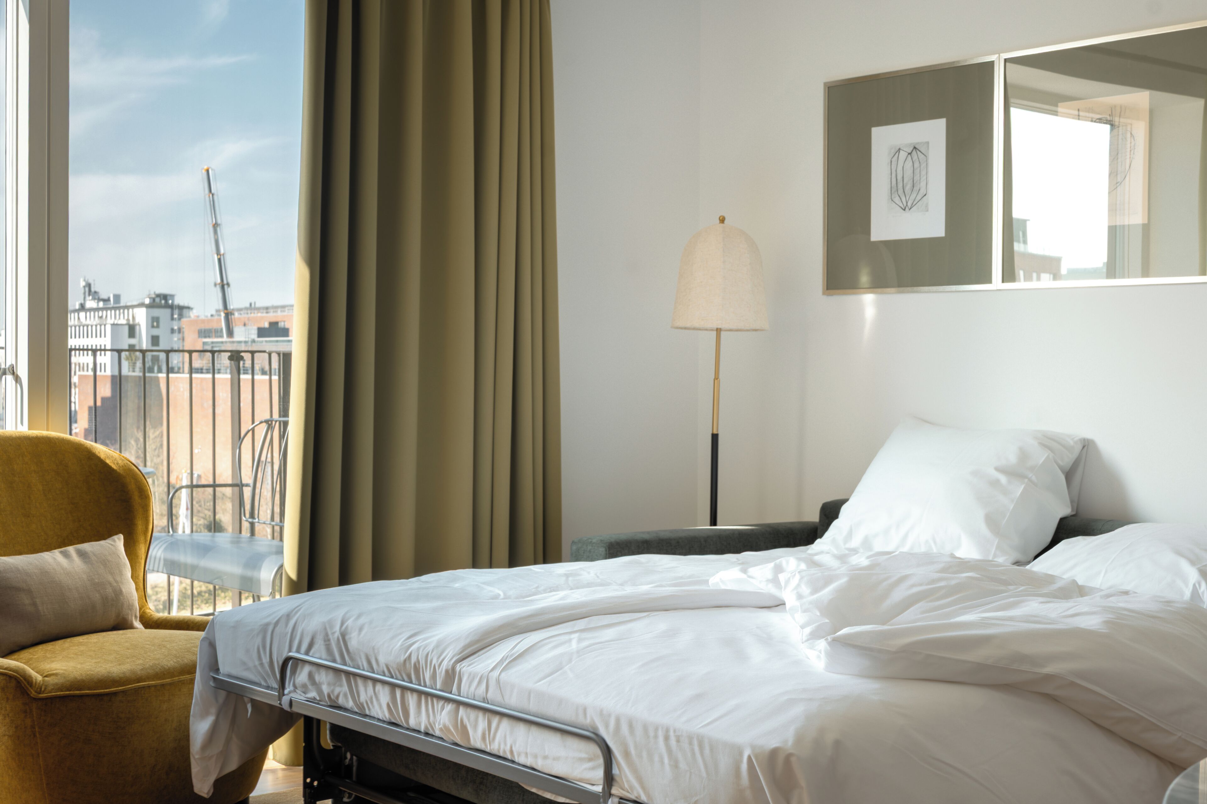 Habitación de hotel con una cama, una silla, una lámpara y vistas a la ciudad a través de una ventana.