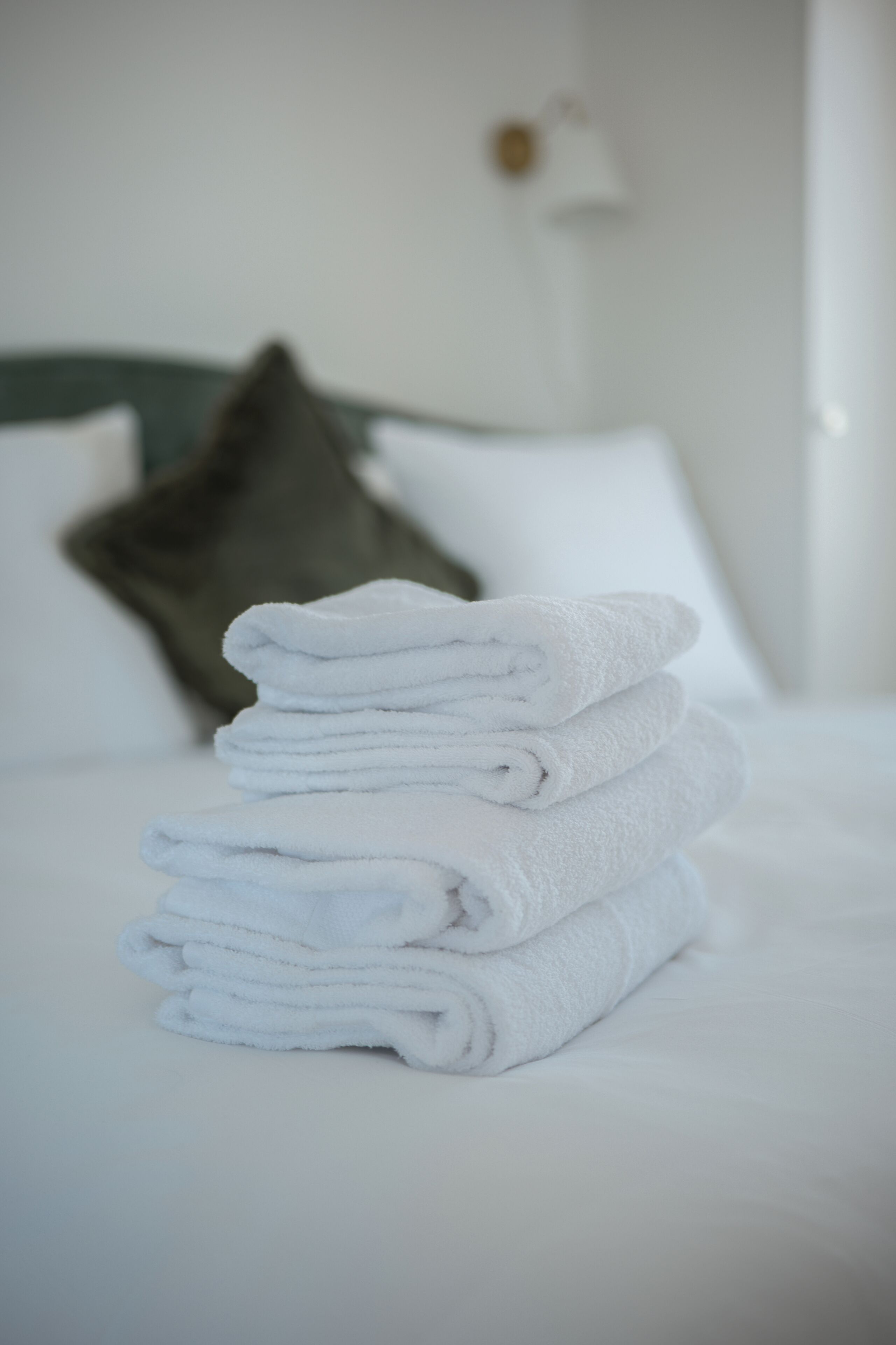 Un montón de toallas blancas ordenadas sobre una cama con ropa de cama blanca y una almohada verde al fondo.