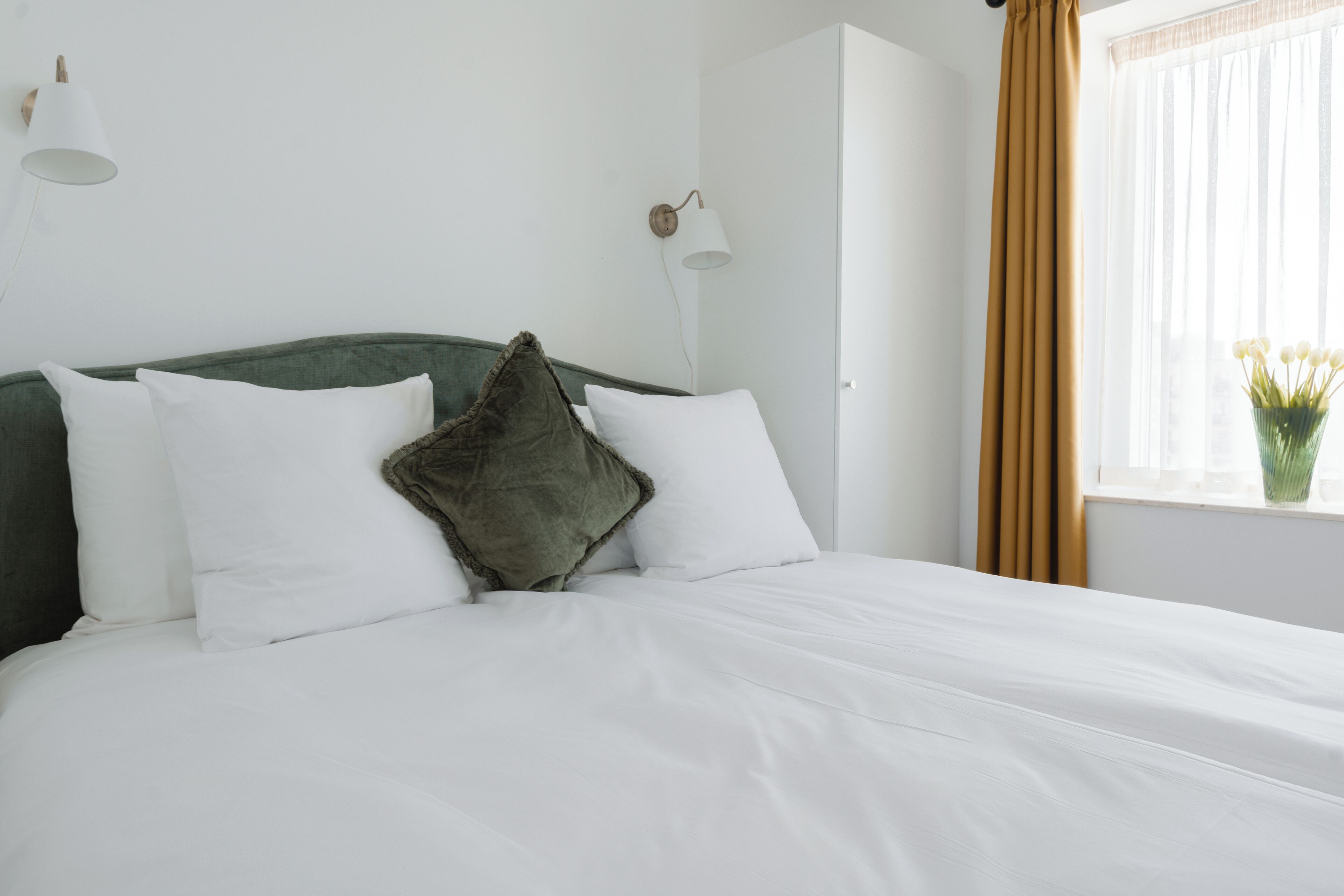 Un lit bien fait, avec du linge de lit blanc et une tête de lit verte, un mur blanc orné d’un luminaire et une fenêtre avec des rideaux.