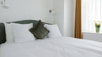Un lit bien fait, avec du linge de lit blanc et une tête de lit verte, un mur blanc orné d’un luminaire et une fenêtre avec des rideaux.
