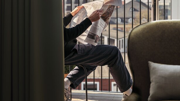 A person reading a newspaper on a balcony with a view of buildings and a church spire. - Clover House by Daniel&Jacob's (Copenhagen)