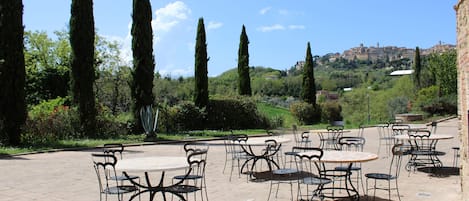 Un patio al aire libre con mesas y sillas redondas, rodeado de cipreses y vistas a un pueblo distante.