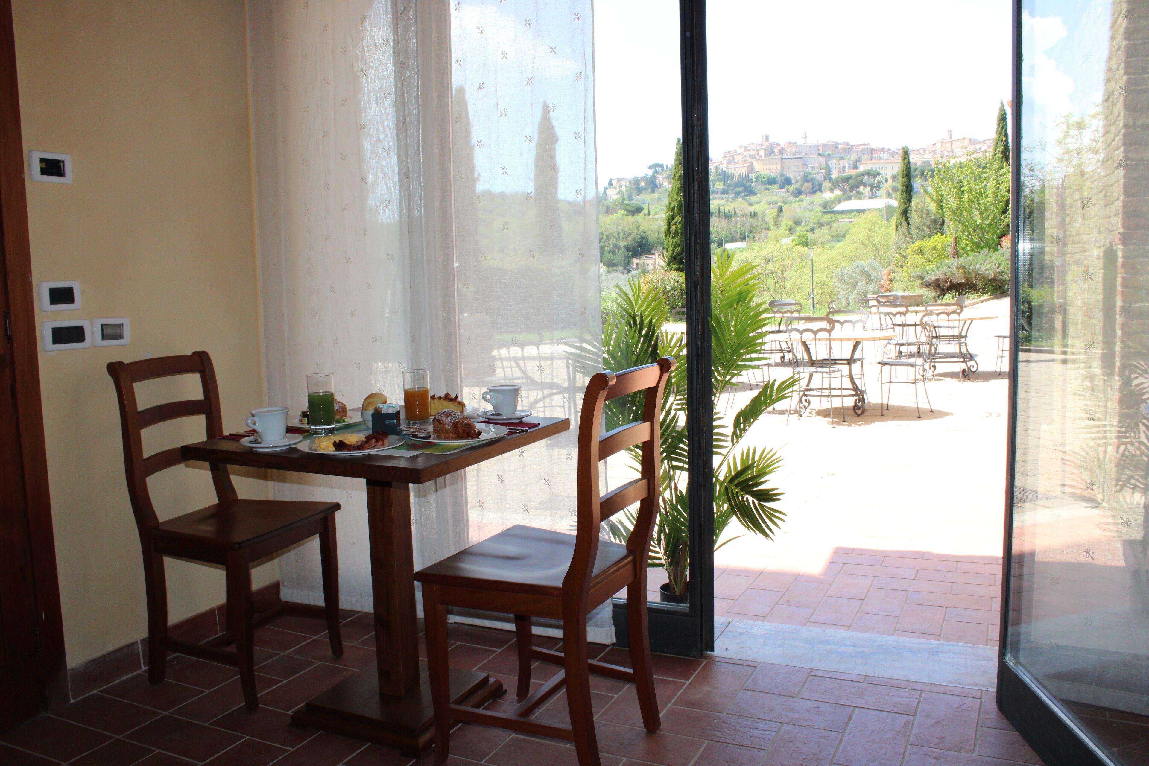 A dining area with a table set for two, chairs, and a view of a garden and cityscape through a glass door.