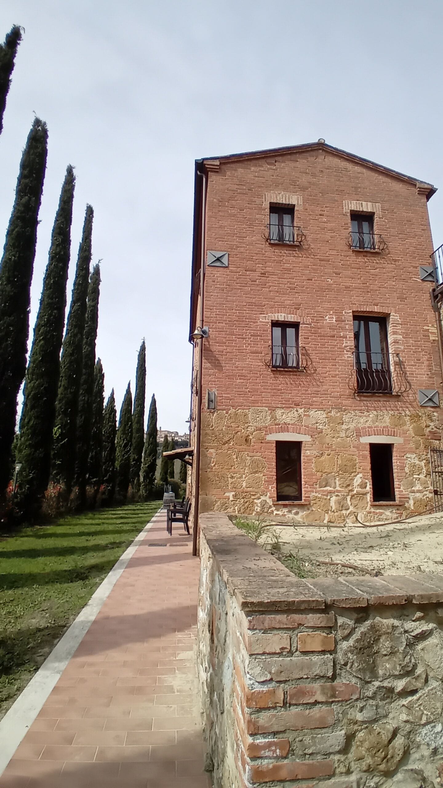 A two-story brick building with multiple windows and balconies, a paved pathway, and tall cypress trees in the background.