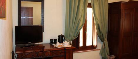 A bedroom with a bed, a television on a wooden dresser, a window with green curtains, and a wooden wardrobe.