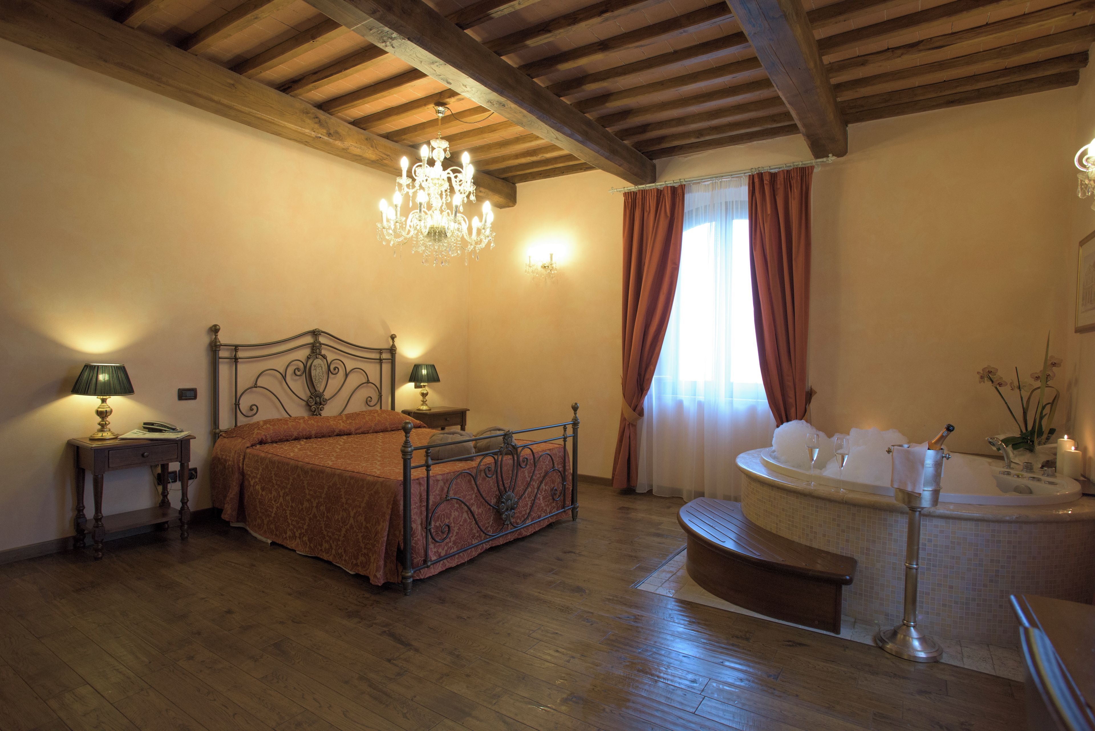 A bedroom with a wooden ceiling, a bed with a metal frame, a chandelier, a nightstand, a lamp, a window with curtains, and a bathroom with a bathtub.