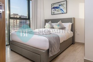 A modern bedroom with a grey bed, white bedding, and a city view through large windows.
