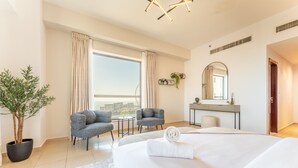 Een moderne hotelkamer met een groot bed, twee fauteuils, een tafeltje, een potplant, uitzicht over de stad en een badkamer met spiegel en wastafel.