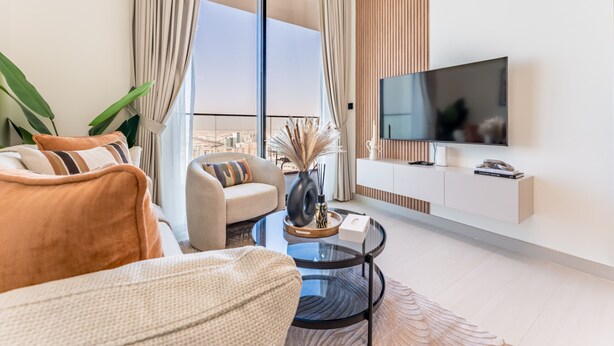 Un salon moderne avec un canapé, un fauteuil et une table basse. Un téléviseur à écran plat est fixé au mur, et une grande fenêtre offre une vue sur la ville.