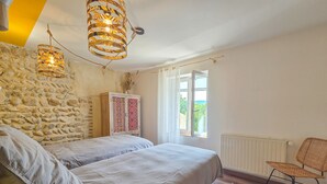 A bedroom with a stone wall, a bed with two pillows, a wooden cabinet, a wicker chair, and a window with a view.