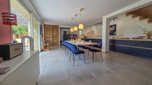 A modern kitchen with a dining area, featuring a large island, pendant lights, and a view through a glass door.