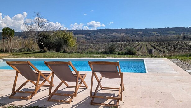 A pool with three wooden lounge chairs facing the water, surrounded by a paved patio.