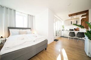 A modern bedroom with a large bed, a desk, and a kitchen area.