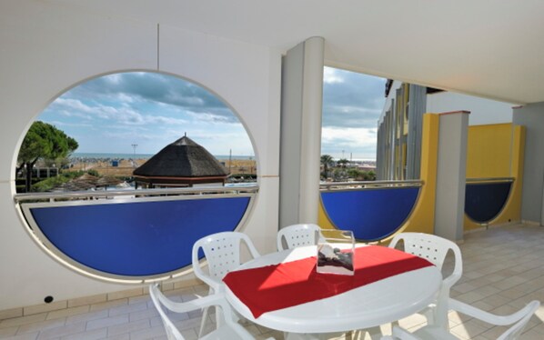 A room with a round table and chairs, a red tablecloth, and a view of a beach with a thatched-roof structure.