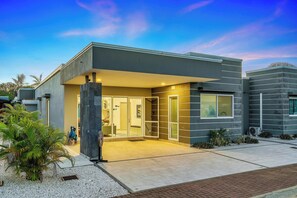Stunning building with a flat roof, glass door, and covered porch area, surrounded by a lush garden with a palm tree.