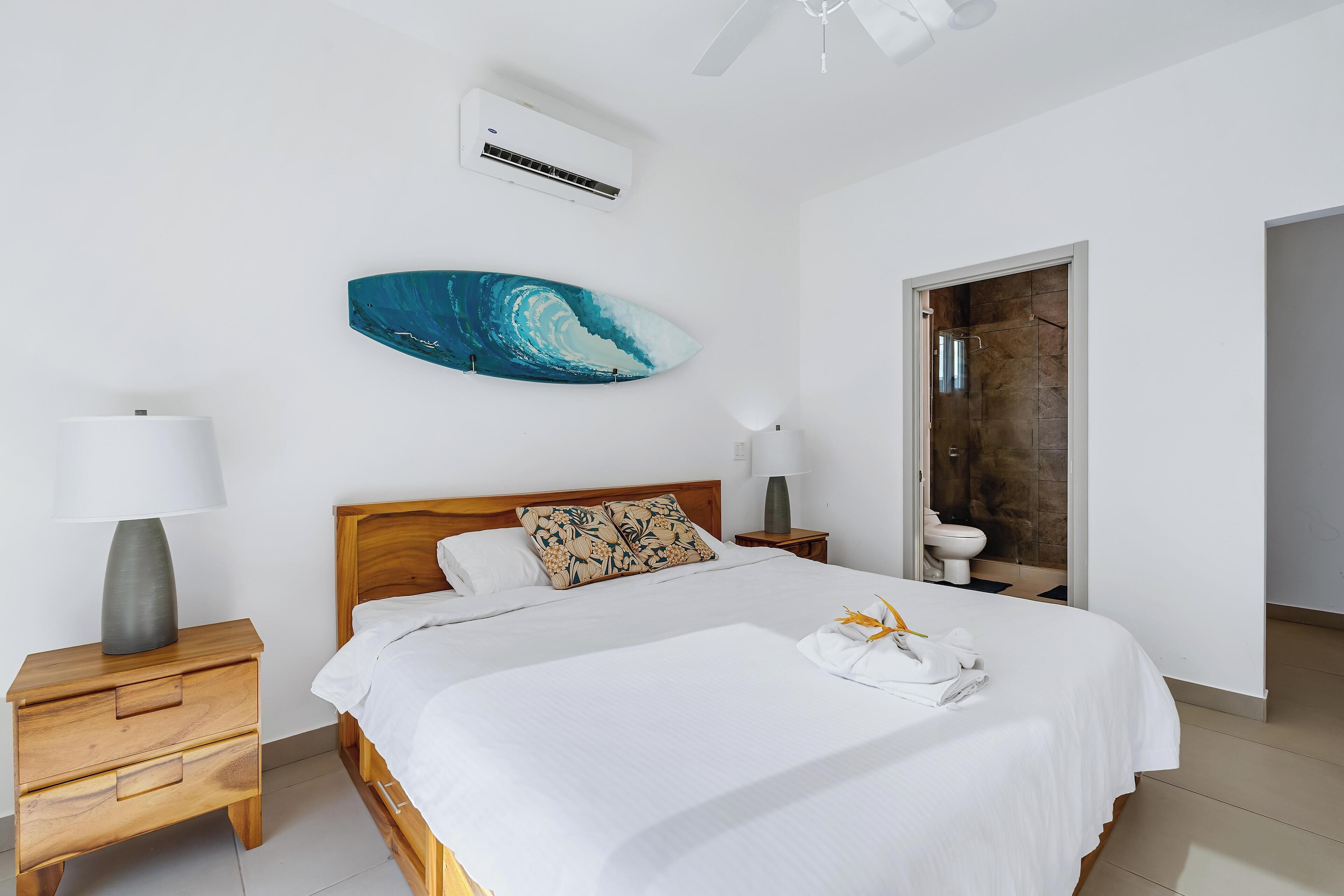 A bedroom with a bed, a nightstand, a wall-mounted air conditioner, a ceiling fan, and a bathroom door.