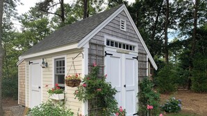 A small, single-story shed with a white door and a window, surrounded by greenery and flowers. - Coastal home in Brewster near beaches and bike path RPMS161 (Brewtser)