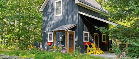A rustic cabin with a large porch, surrounded by a garden and a stone wall.