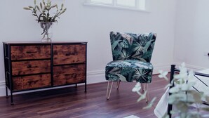 A room with a wooden cabinet, a chair with a tropical print, a table, and a window. - Converted bank: centre of Town (split apartment) (Holmfirth)