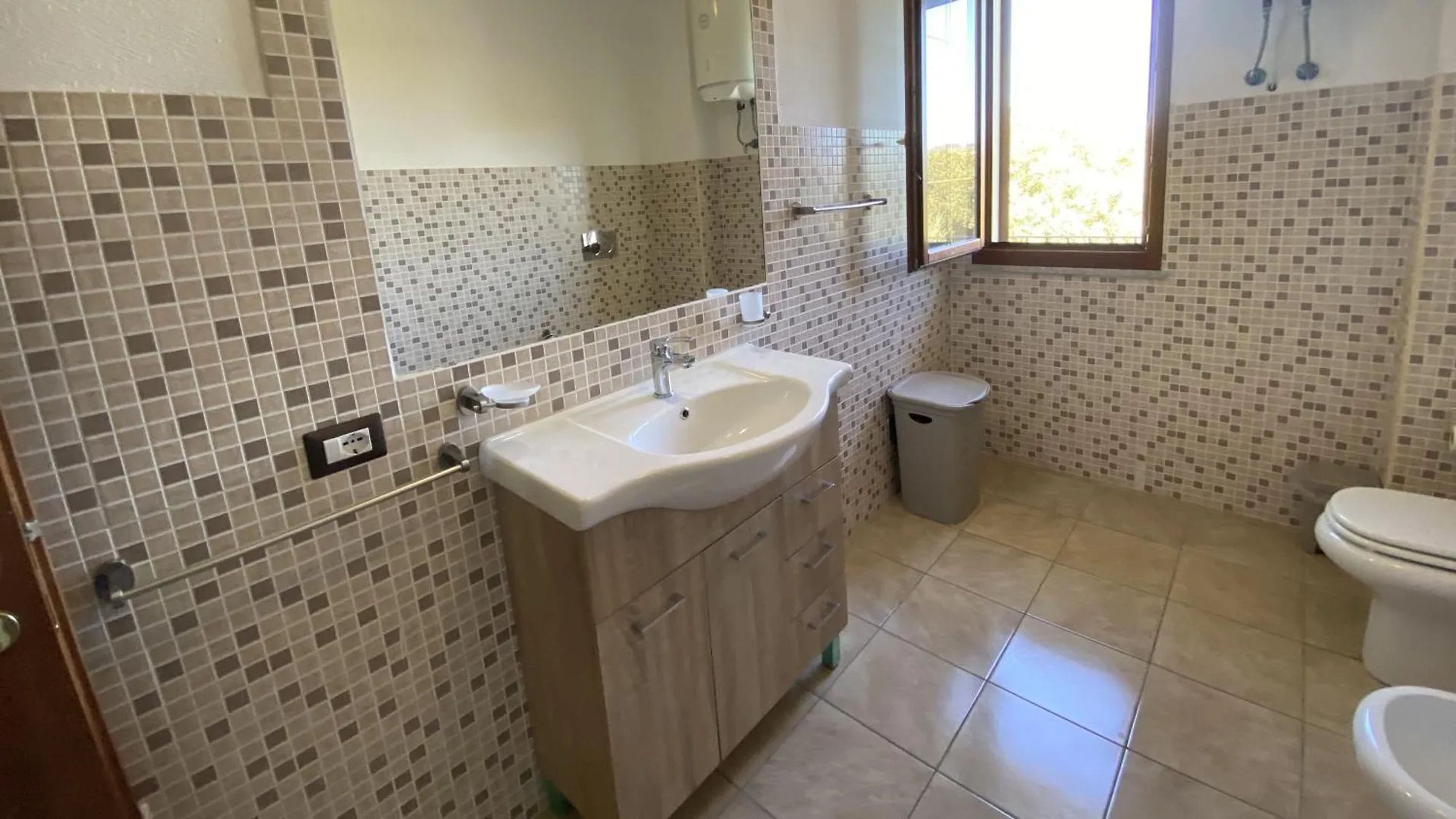 A bathroom with a tiled floor, a sink with a mirror, a toilet, and a window.