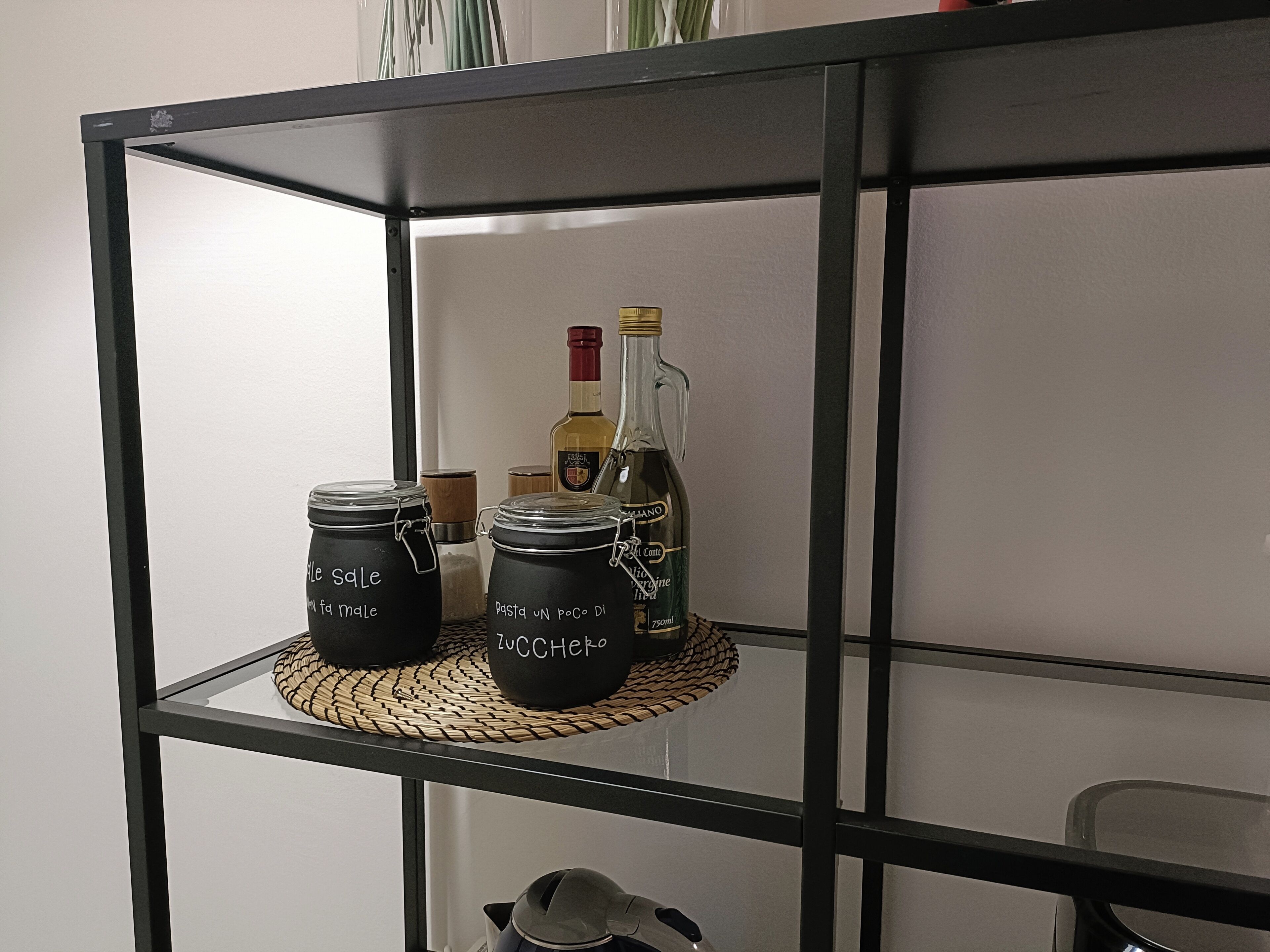 Ein schwarzes Metallregal mit Gläsern, beschriftet mit „Sale“ und „Pasta un poco di zucchero“, auf einer gewebten Matte.