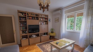 Una sala de estar con una estantería de madera, un televisor, una mesita con un tapete y un ventanal con cortinas.