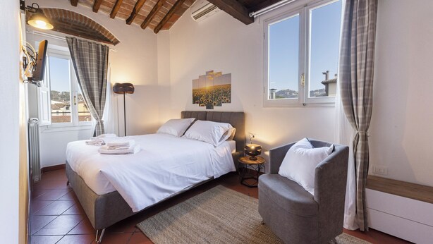 A bedroom with a bed, a grey armchair, a bedside table, and a window with curtains. - Yellow Home by MMega (Florence)