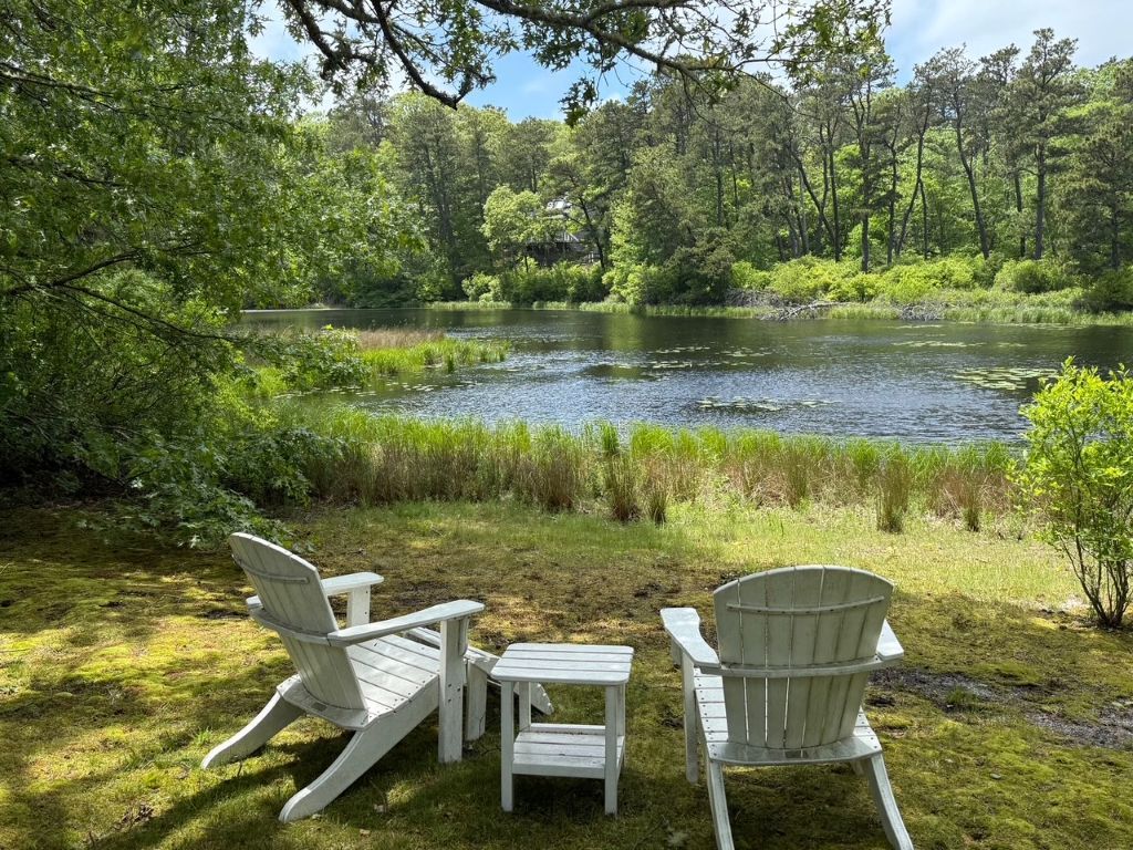 A lakeside setting with three Adirondack chairs and a small table, surrounded by lush greenery and trees.